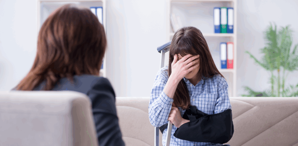 Injured woman with an arm sling and crutches sitting on a couch, covering her face while speaking with another woman