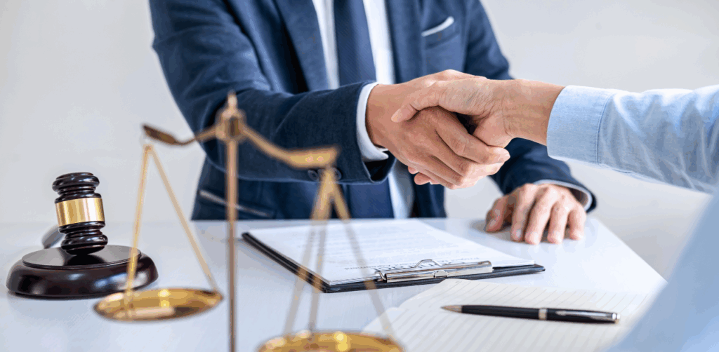 Lawyer in a dark suit shakes hands with a client across a desk featuring legal documents, a gavel, and justice scales—symbolizing a successful personal injury settlement or legal agreement