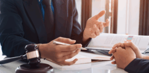 Lawyer in a suit gestures while advising a client at a desk covered with legal documents and a gavel, suggesting a personal injury or car accident case consultation