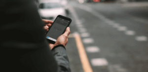 Man standing by the roadside holds a smartphone showing a rideshare app with a map and pickup location, waiting for his driver to arrive