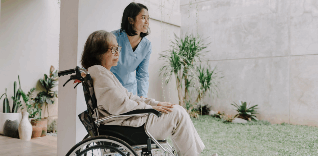 Smiling caregiver gently pushes an older woman in a wheelchair through a bright, peaceful courtyard, illustrating compassionate elder care in a supportive environment