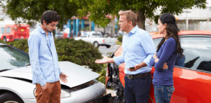 Two men argue next to damaged cars after a crash, with a woman standing by listening