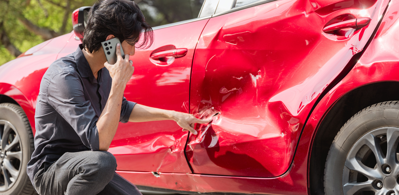 Man on phone kneels beside red car, inspecting large dent from a side collision