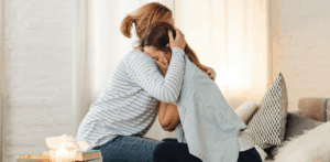 Two women embrace in a comforting hug, one visibly emotional on a living room couch