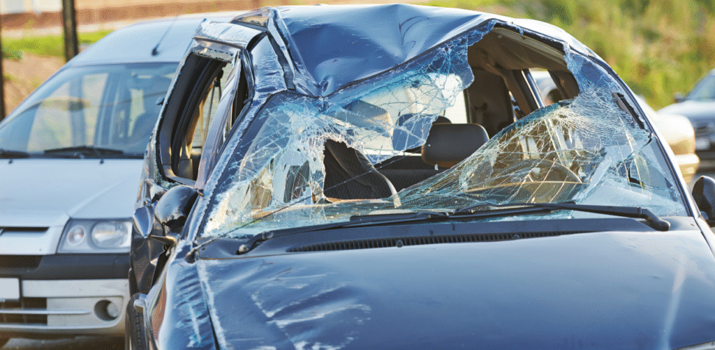 Severely damaged car with shattered windshield and crushed roof after traffic accident, another vehicle stopped behind