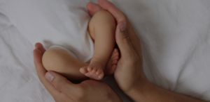 Adult hands gently holding a baby’s legs and feet while the infant lies on a soft white blanket