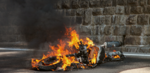 Motorcycle engulfed in flames on a roadway beside a stone wall, with thick black smoke rising into the air.