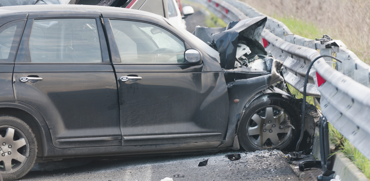 Severely damaged car with front-end impact crashed into a highway guardrail, showing heavy collision damage and debris on the road