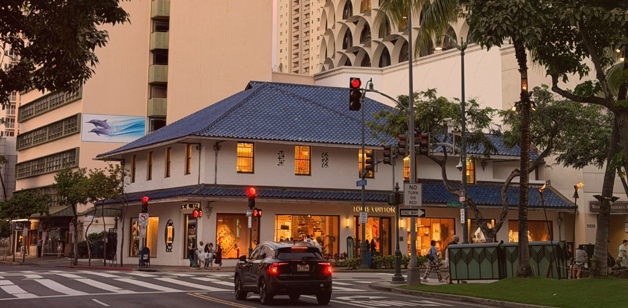 Urban street intersection at dusk with cars stopped at red lights beside storefronts and multi-story buildings