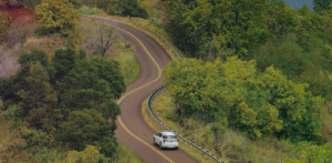 White SUV drives along a winding two-lane road through dense green forest and hillside vegetation